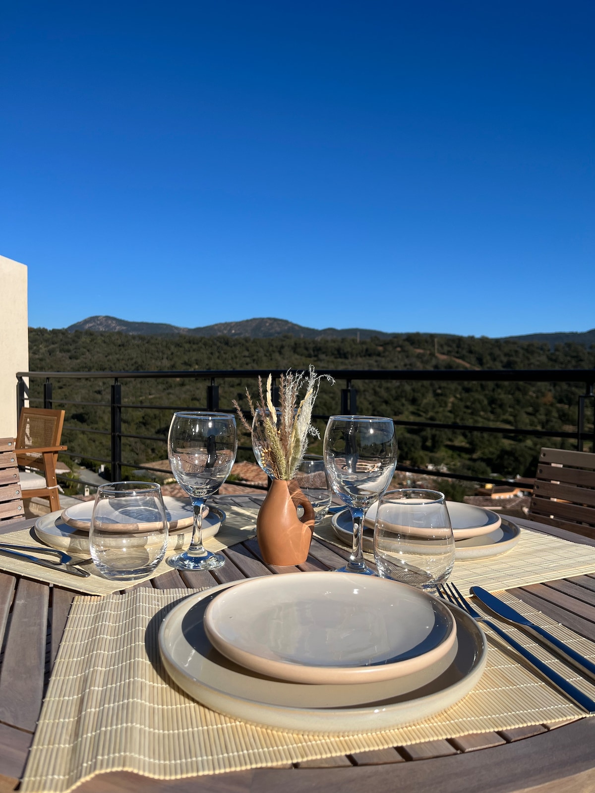 A dining table is set on a terrace, featuring plates, glasses, and a small vase with dried flowers. The natural landscape of mountains is visible in the background, under a clear blue sky.