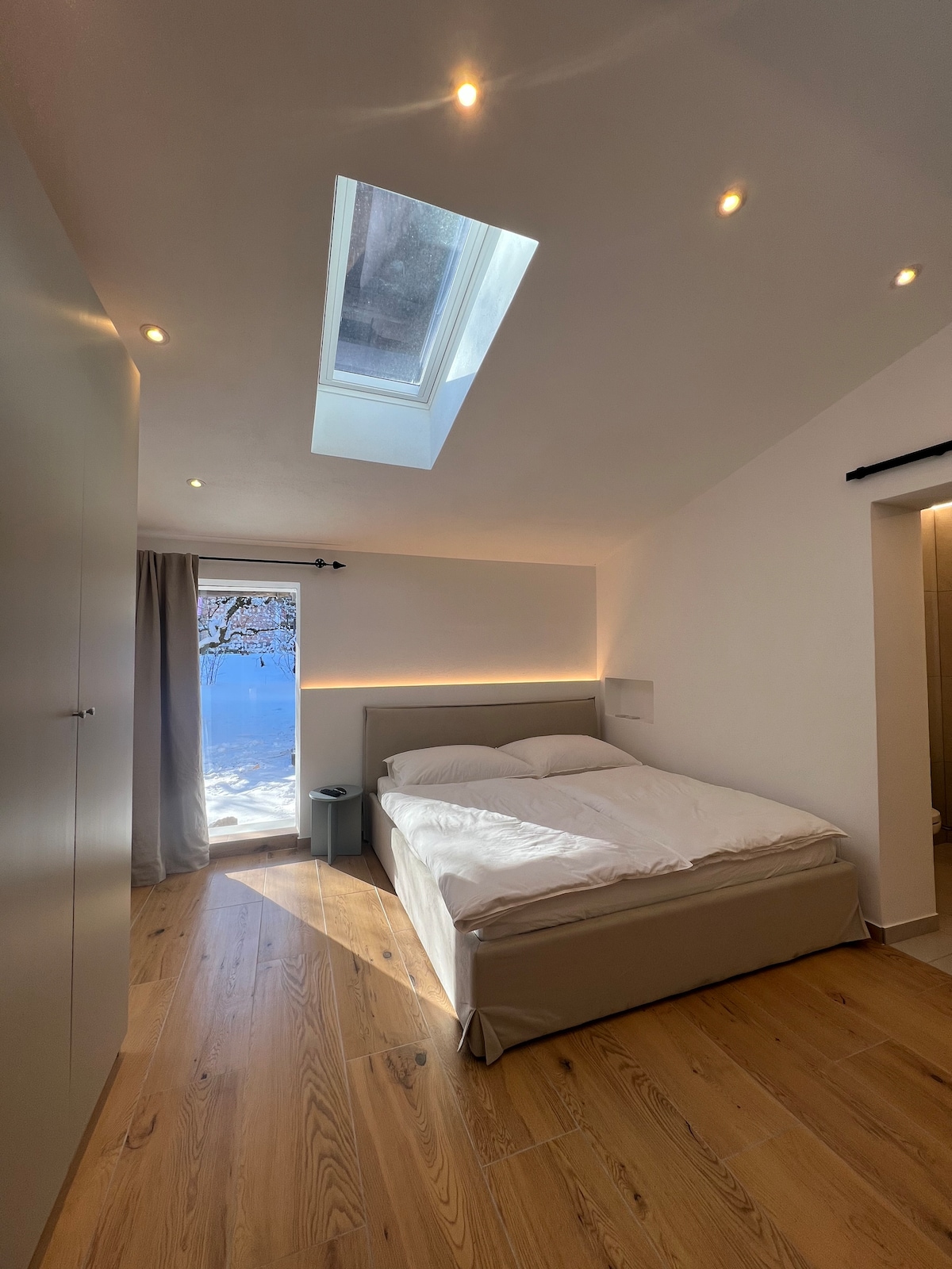 A spacious bedroom features a large bed with clean white linens, under a skylight that allows natural light to enter. Warm wood flooring complements the minimalistic design. A doorway leads to an outdoor view of snow-covered landscape, enhancing the serene atmosphere.