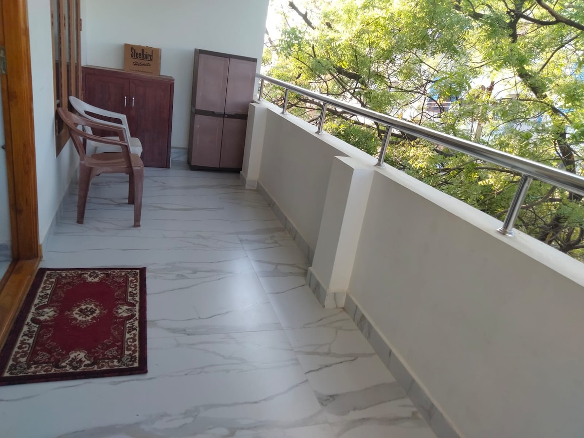 A balcony space features a smooth tiled floor, complemented by a decorative area rug. Two wooden chairs are positioned for seating, and a small cabinet is visible nearby. Lush greenery is seen through the railing, providing a calming view.