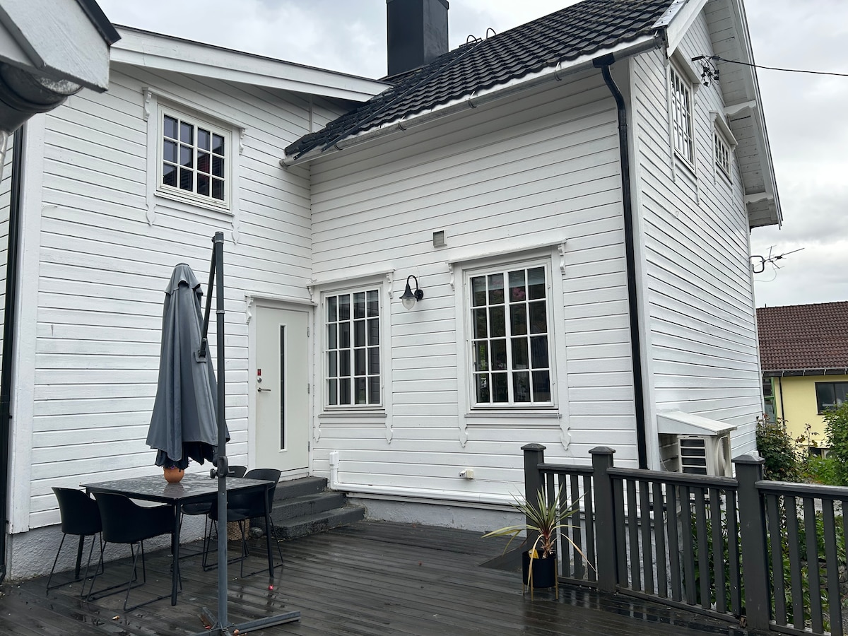 The exterior of a white wooden house is shown, featuring large windows that allow natural light to illuminate the space. A deck area is visible, furnished with a table and chairs, alongside a grey umbrella. The entrance door is centered, framed by classic architecture.