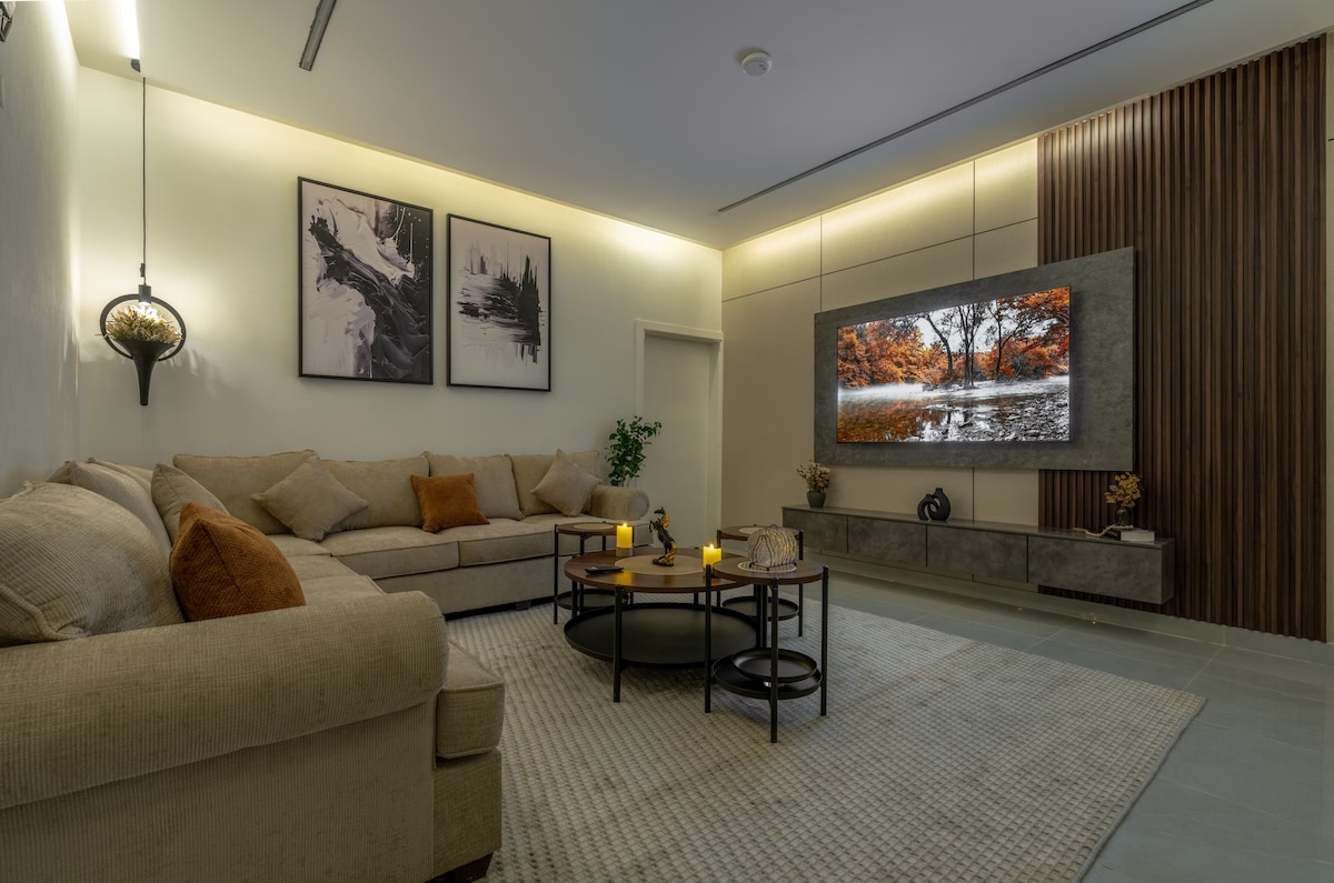 A spacious living area features a large beige sectional sofa adorned with orange accent pillows. A round coffee table sits in the center, surrounded by a light-colored area rug. A large television is mounted on the wall, complemented by two framed artworks and soft ambient lighting.
