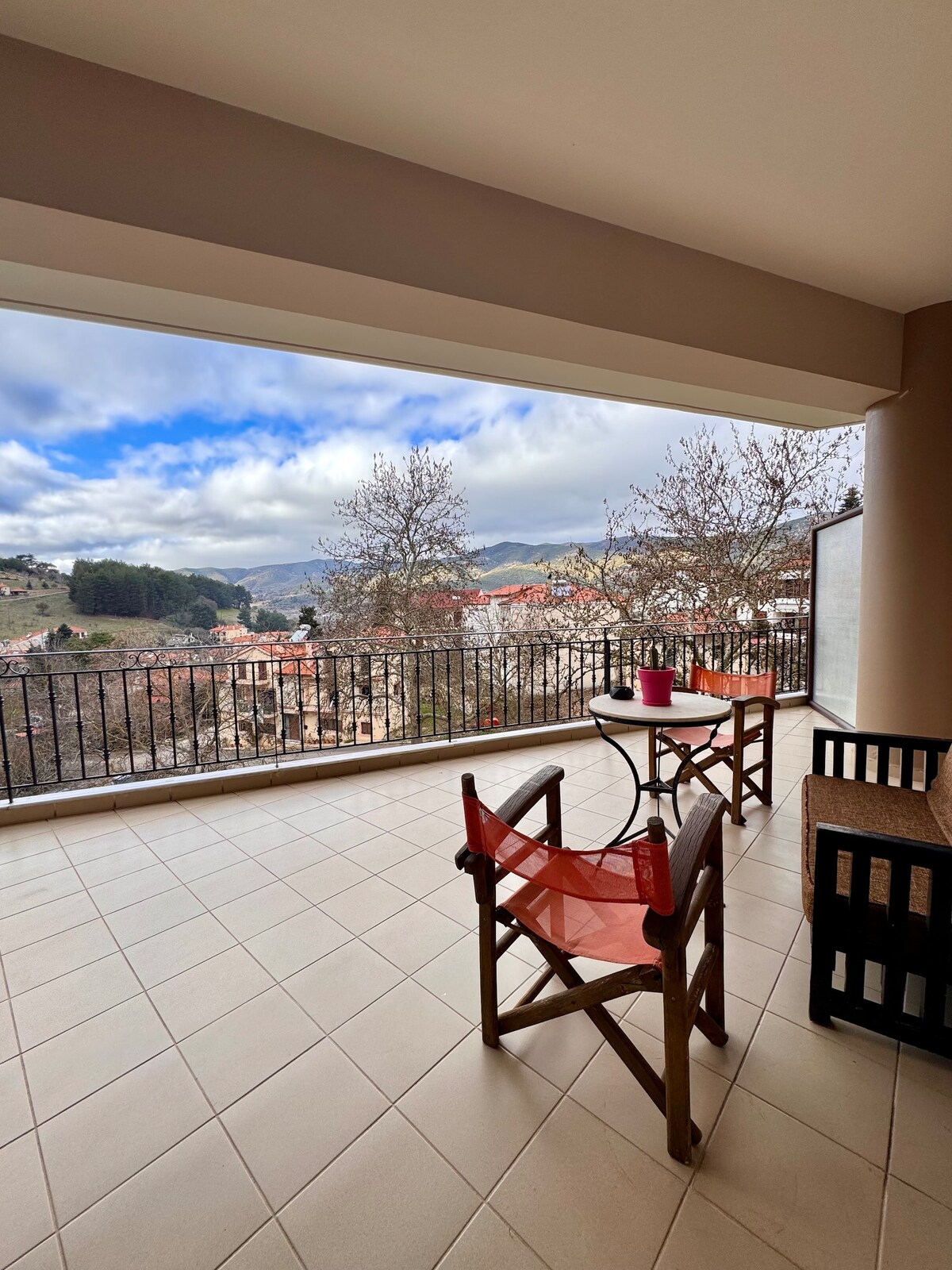 A spacious balcony offers stunning views of the surrounding landscape. Two wooden chairs are positioned around a small table, ideal for outdoor relaxation. The area is framed by a protective railing, and the tiled floor complements the serene environment.