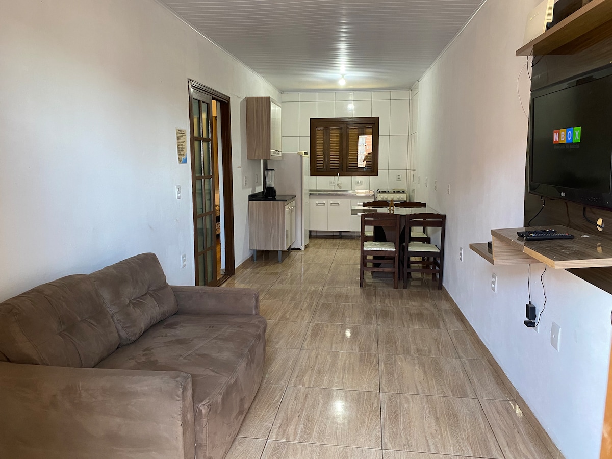 A shared living space features a comfortable sofa and a dining area with a wooden table and chairs. The kitchen is visible in the background, equipped with cabinetry and essential appliances. Natural light enters through a window, illuminating the tiled floor.