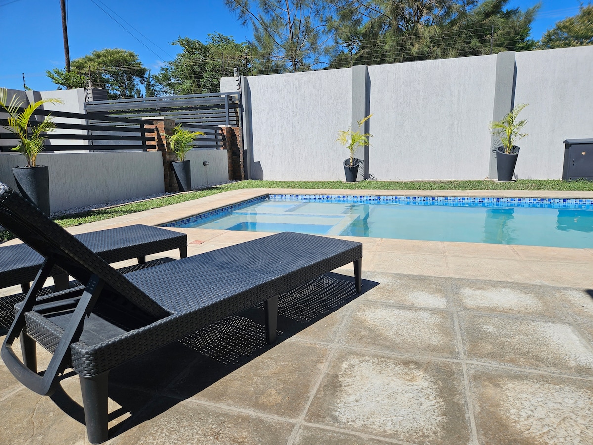 A swimming pool is framed by a tiled patio, featuring two lounge chairs positioned nearby. Green plants in black pots add a touch of greenery, while a low wall provides privacy. Clear blue water reflects the sunlight, creating a relaxing outdoor space.