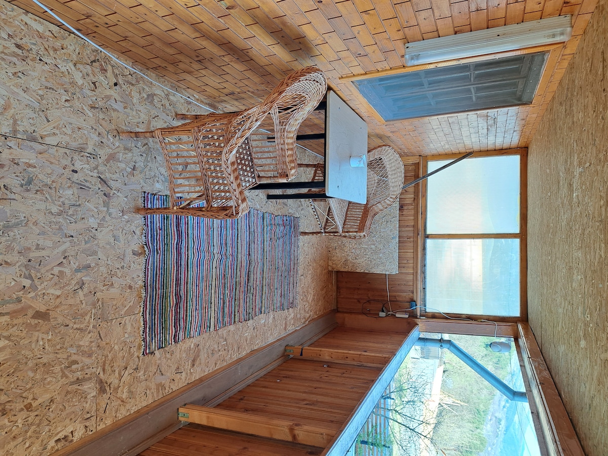A sunlit sunroom features a wooden floor and walls lined with natural wood paneling. Two woven chairs are positioned around a small table, accompanied by a colorful striped rug. Large windows provide a bright view of the outdoors.