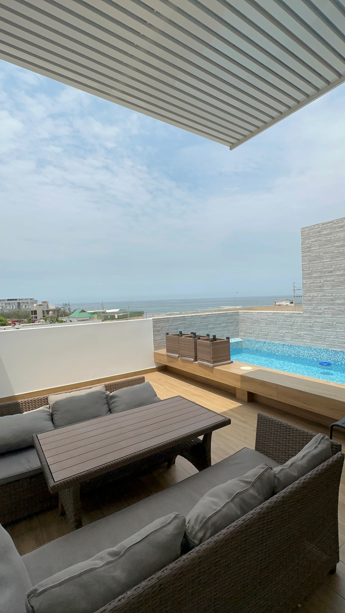 An outdoor terrace is featured with a comfortable seating arrangement, including a sectional sofa and a coffee table. A small pool with clear blue water is visible, surrounded by a stone wall. The horizon showcases glimpses of the sea, framed by soft cloud cover.