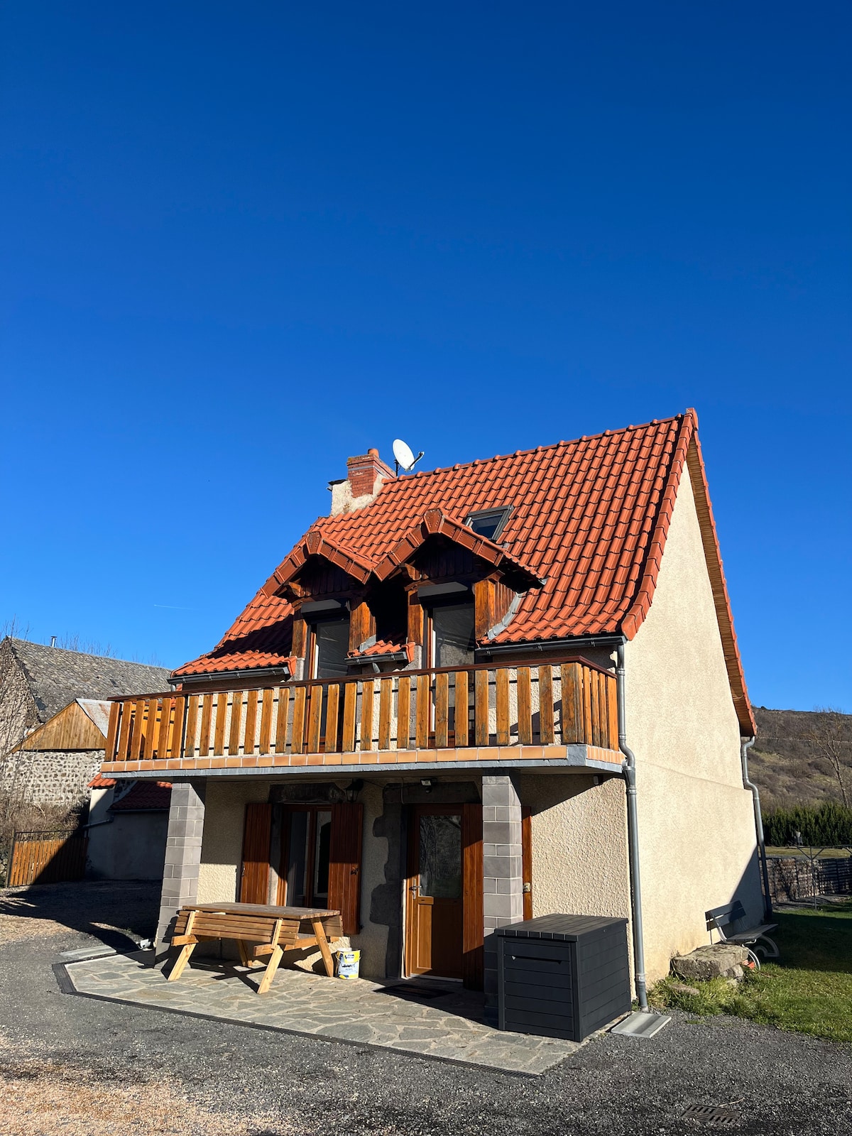 The front exterior of a charming two-story house features a sloped red tile roof and brown wooden accents. A balcony is accessible from the upper floor, and a stone pathway leads to the entrance. Clear blue skies provide a backdrop for the home.
