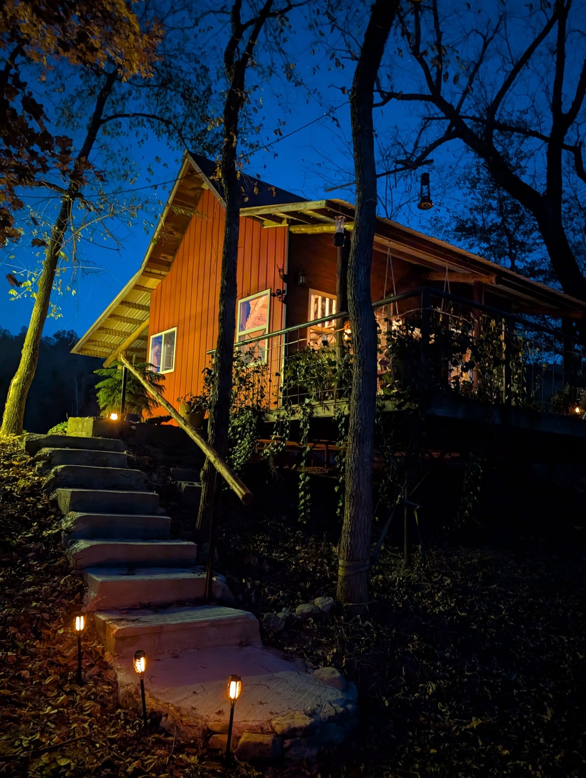 The cabin stands amidst trees, illuminated by soft exterior lighting. Stone steps lead up to the entrance, bordered by ground lights. The rustic, red exterior is complemented by a covered porch, inviting evening relaxation in the tranquil setting of the Ozarks.