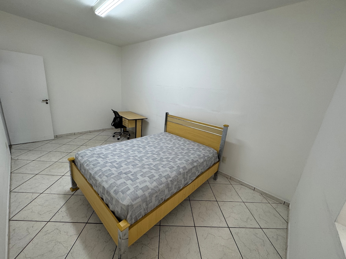 A simple bedroom features a light-colored wooden bed with a patterned grey mattress, positioned against a white wall. Natural light enters through a window. A desk with a chair is located beside the bed, and the floor is covered with light-colored tiles.