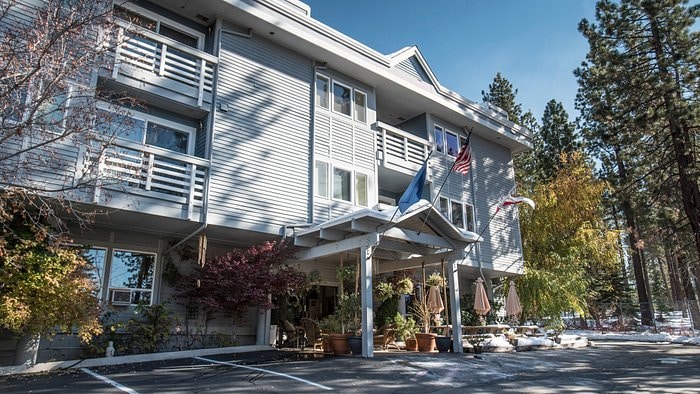 The exterior of a three-story building features a porous façade with large windows. A covered entrance area includes potted plants and flags, while surrounding trees and a clear sky enhance the sense of space. A parking area is visible in the foreground.
