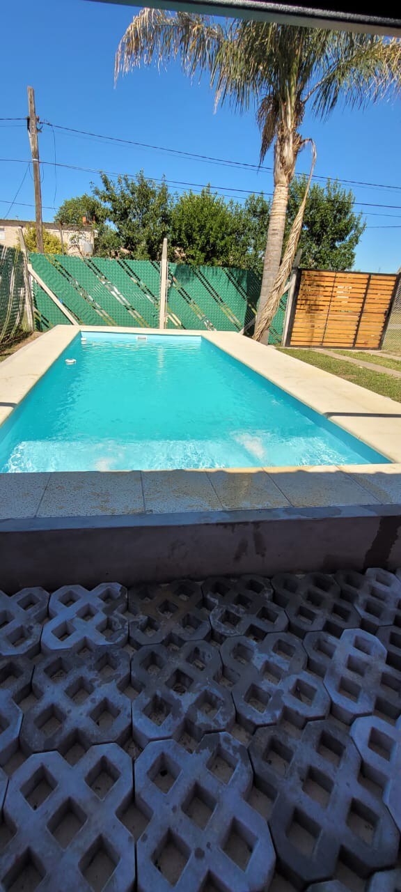 A serene swimming pool is framed by a light-colored deck and lush greenery. The clear blue water reflects sunlight, while a grassy area alongside the pool provides a space for relaxation. Fencing offers privacy, enhancing the inviting outdoor setting.