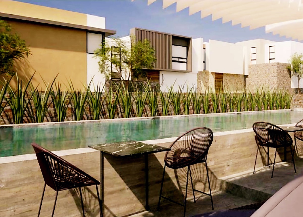 An outdoor area features a tranquil pool bordered by a lush garden of tall plants. Several black wicker chairs surround simple stone tables, creating a relaxed setting for guests to enjoy.