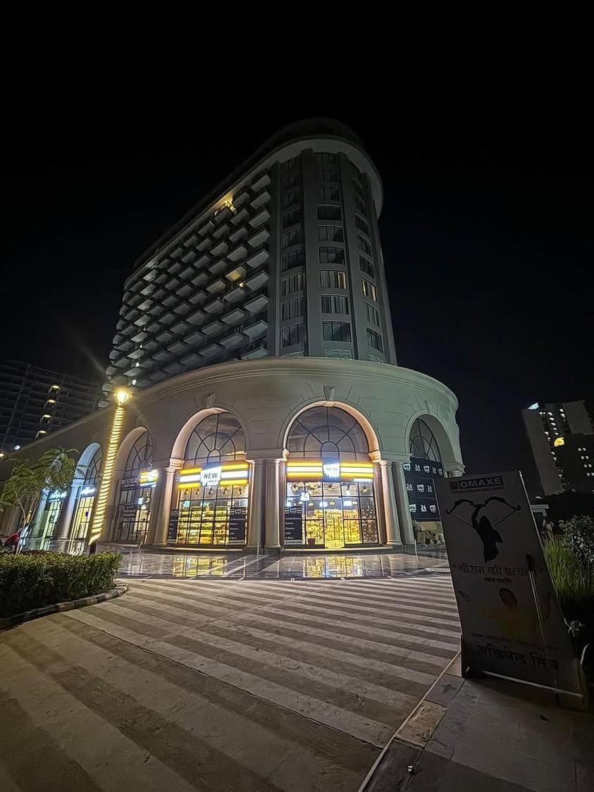 The modern building features a sleek, curved design illuminated against the night sky. Arch-shaped windows showcase warm lights inside, while the entrance is framed by neatly arranged landscaping. The nearby sign and pavement lead towards the structure, highlighting its contemporary style.