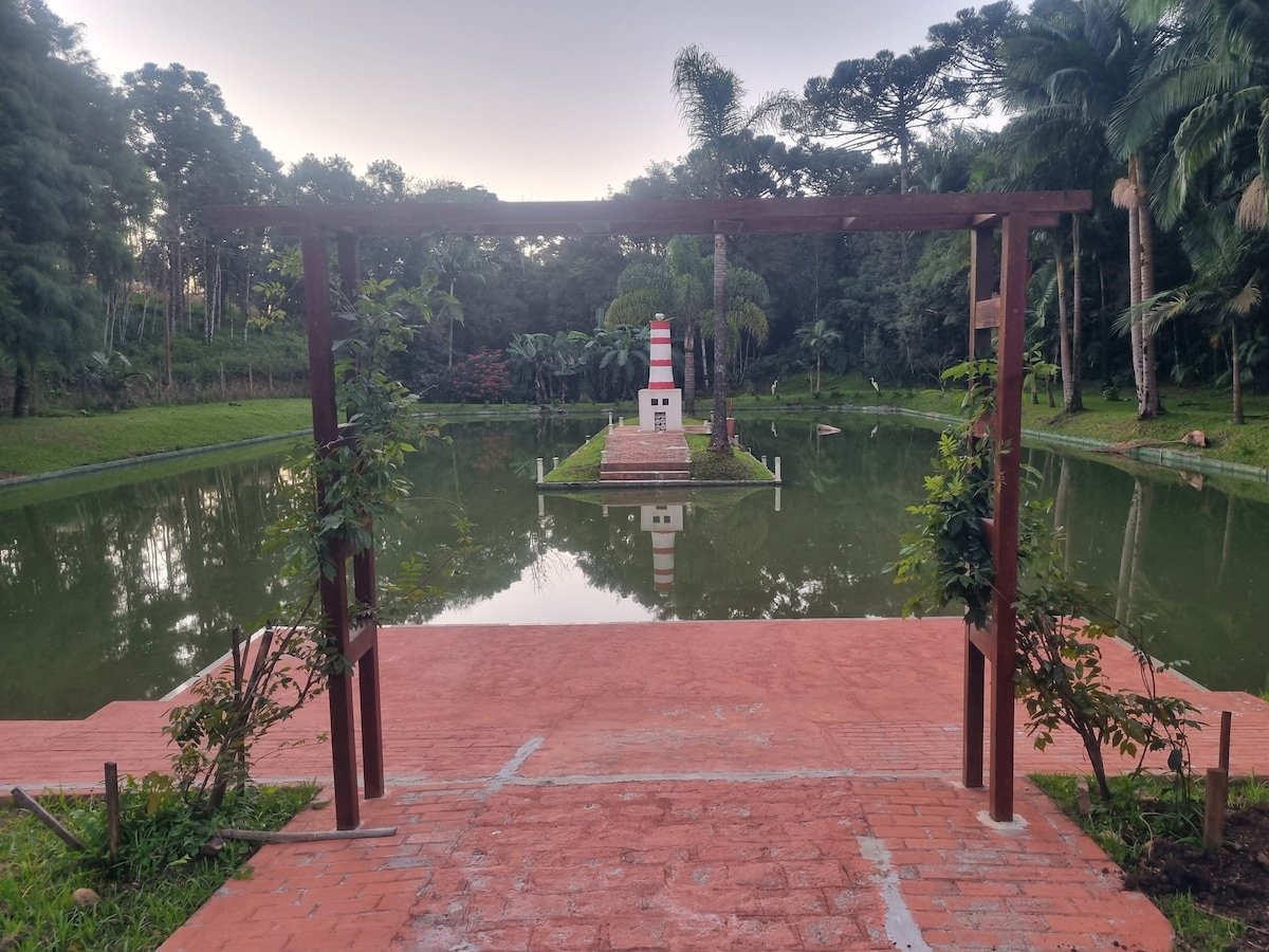 A serene landscape features a peaceful pond surrounded by lush greenery. A small island, highlighted by a slender lighthouse, is visible in the center. A wooden archway adorned with climbing plants frames the view of the water, enhancing the natural setting.