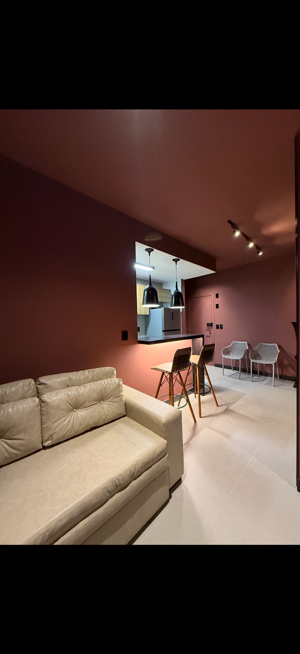 A modern living space features a light-colored sofa positioned on a tiled floor. Two minimalist chairs and a dining table with two stools are visible, accompanied by an open kitchen area that showcases pendant lighting and a dark-colored countertop.