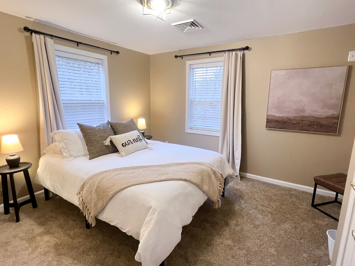 A well-lit bedroom features a spacious bed dressed in crisp white linens, topped with a soft tan throw. Two bedside lamps provide warm illumination, and light-colored curtains frame the windows, enhancing the natural light. A minimalist painting adorns one wall, adding subtle decor.