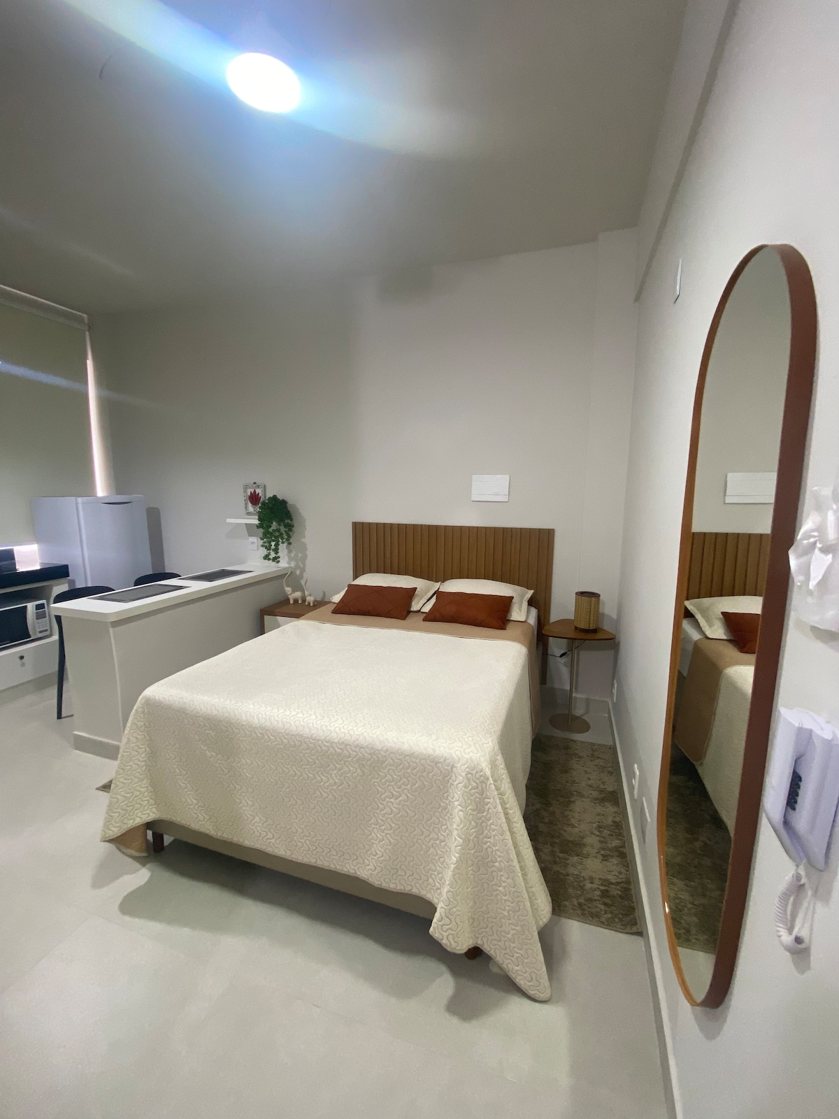 A well-lit room features a spacious bed covered with a light-colored bedspread, accented by two decorative pillows. A mirrored accent is positioned on the wall, reflecting the inviting atmosphere. A small bedside table and kitchenette are also visible, contributing to functionality.