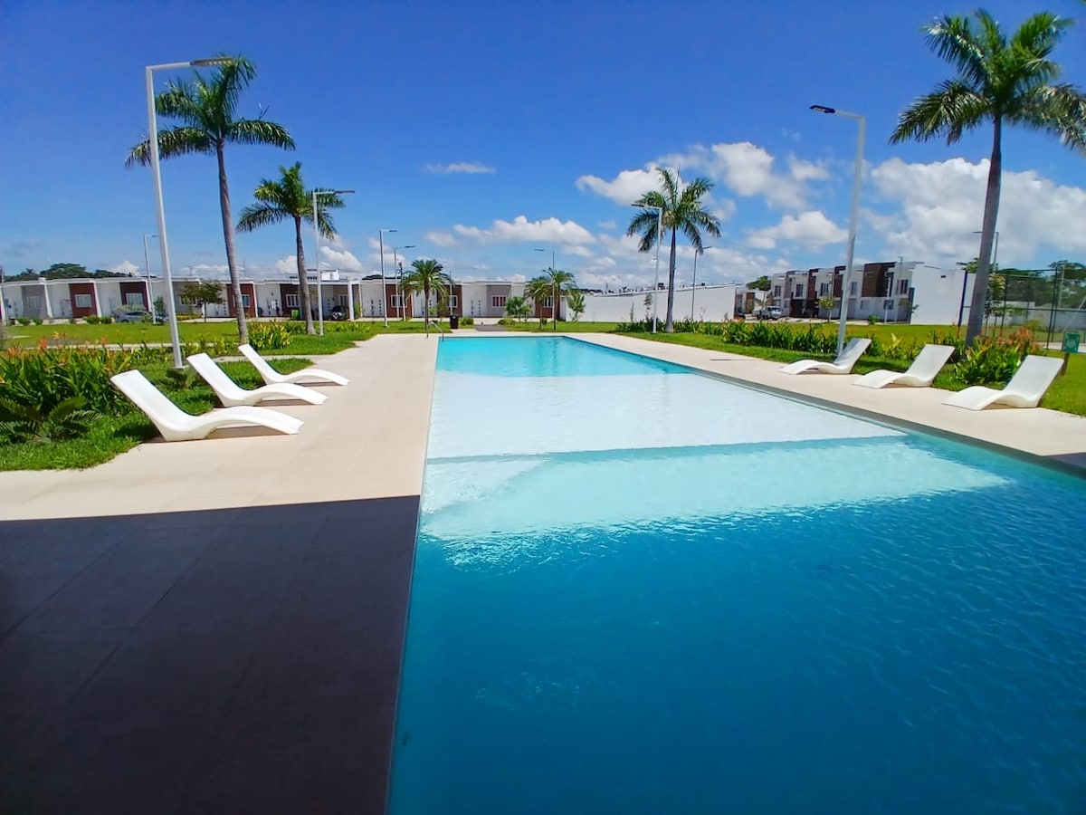 An expansive swimming pool is bordered by lounge chairs, surrounded by landscaped greenery and palm trees. The pool's clear blue water reflects the sky, creating a serene environment in a well-maintained outdoor space.