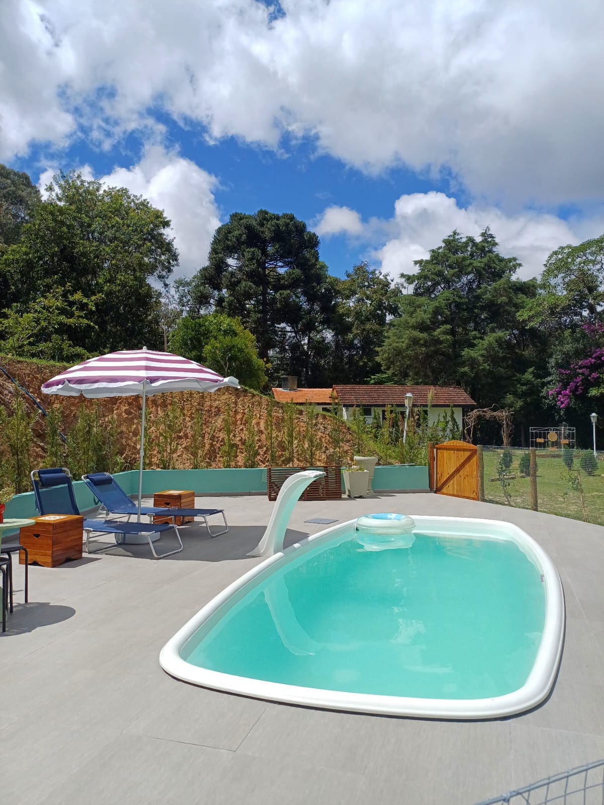A private pool is set in a spacious outdoor area, surrounded by greenery. Lounge chairs and a sun umbrella offer relaxation options, while a wooden gate provides a secure entrance. The bright blue sky, with scattered clouds, enhances the serene atmosphere of the space.
