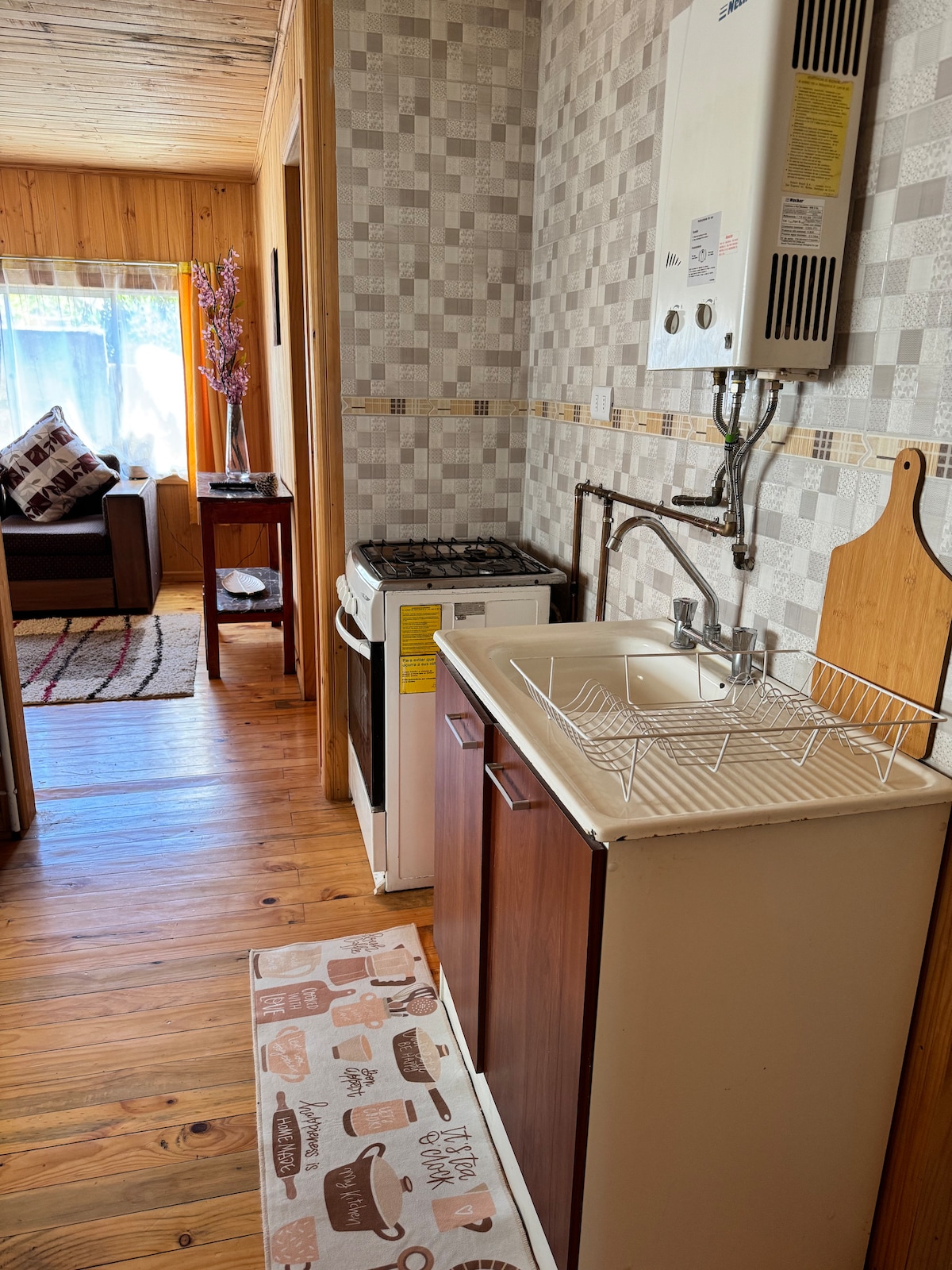 A compact kitchen area features a small sink and a stove with an oven. Light-colored cabinetry complements the tiled backsplash. A kitchen towel sits on the floor, and natural light filters through a nearby window, illuminating the space.