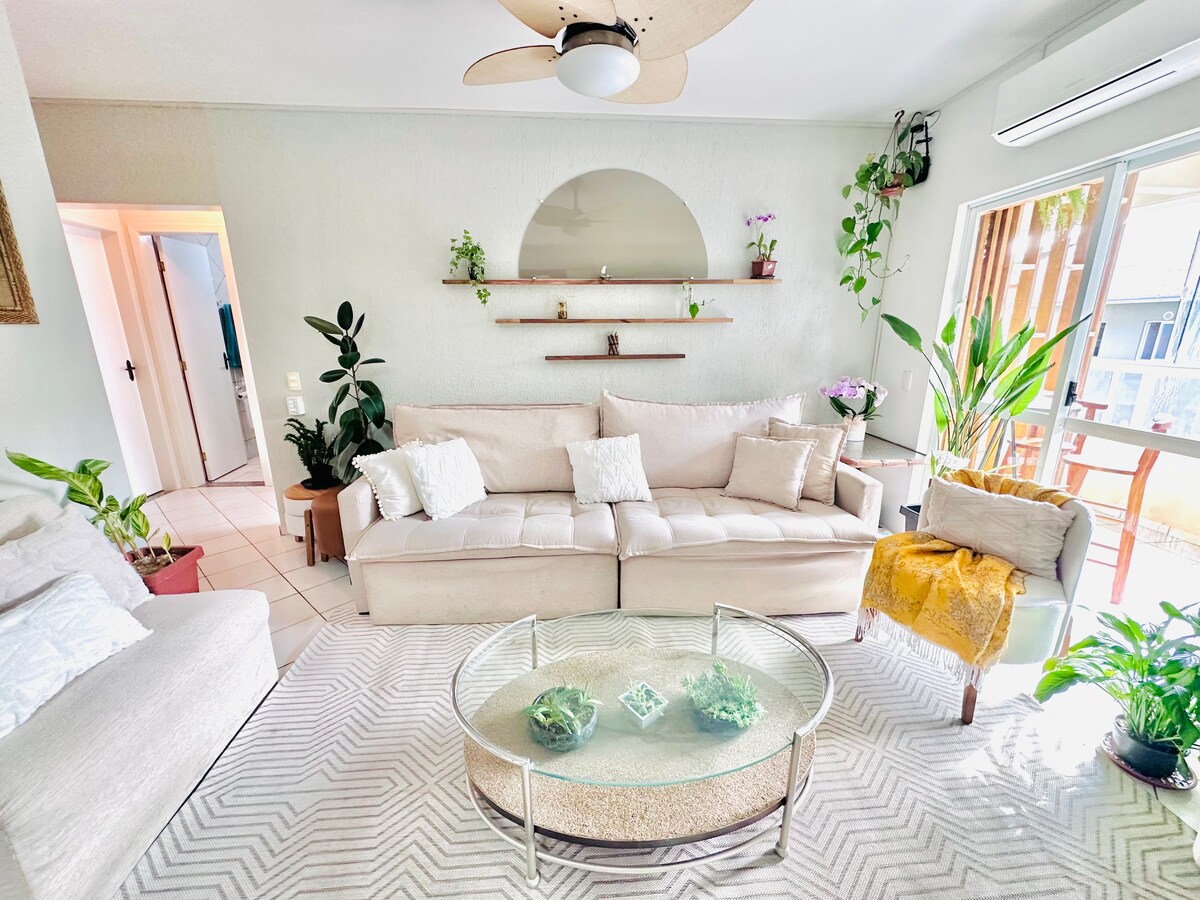 The living area features a light-colored sectional sofa with decorative cushions, accompanied by a cozy armchair. A circular glass coffee table sits on a textured rug. Green plants are displayed around the space, enhancing the natural light from the nearby window.