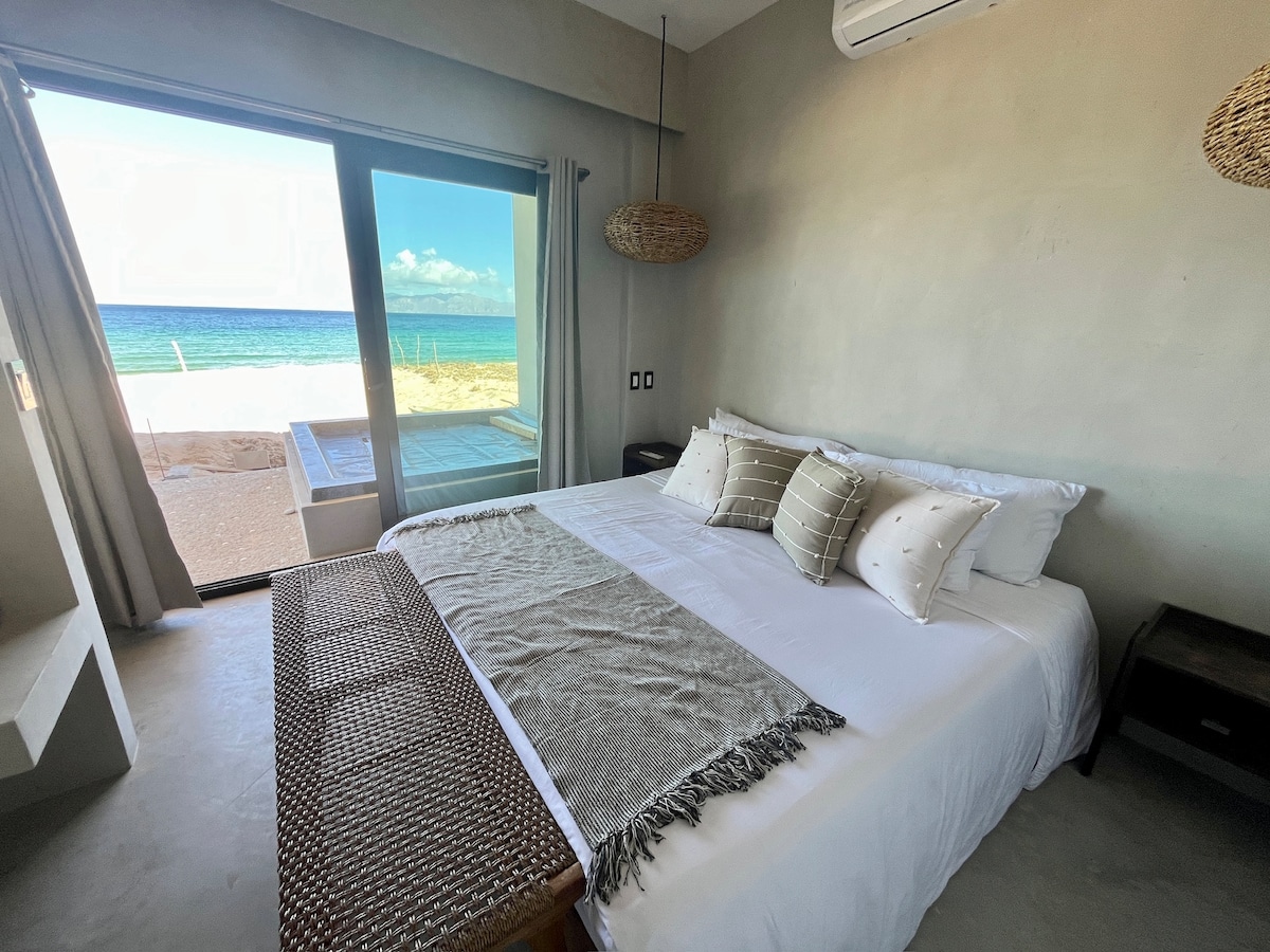 A spacious bed with neutral linens and decorative pillows faces large sliding glass doors that lead to a private outdoor area. Natural light floods the room, highlighting the minimalist decor and coastal views beyond.