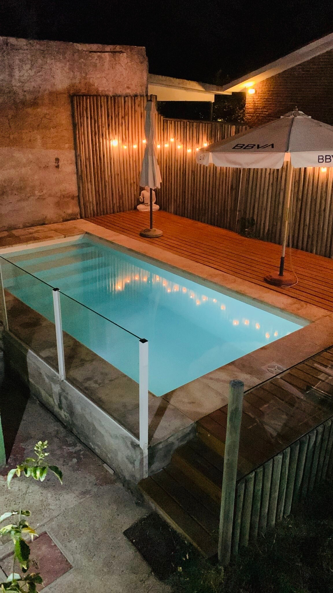 A well-lit, private pool area is displayed at night, featuring a clear pool surrounded by wooden decking. Soft string lights hang overhead, creating a serene ambiance. A patio umbrella stands at one corner, enhancing the outdoor space's inviting nature.