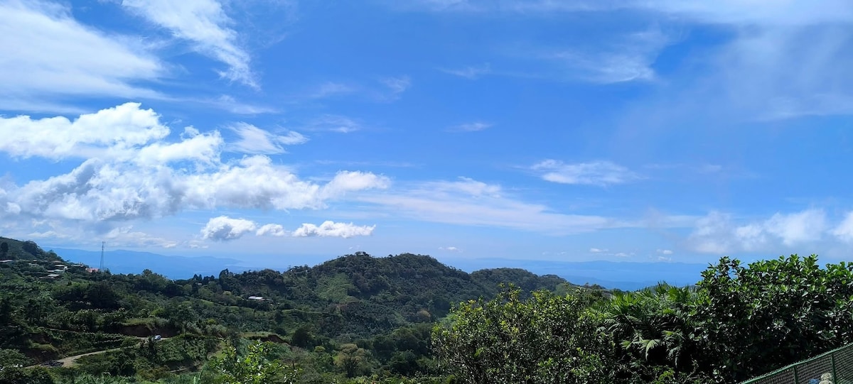A panoramic view of the surrounding mountains features rolling hills marked by lush greenery under a bright blue sky with scattered clouds. The horizon extends towards the distant mountains, creating a serene and expansive landscape.
