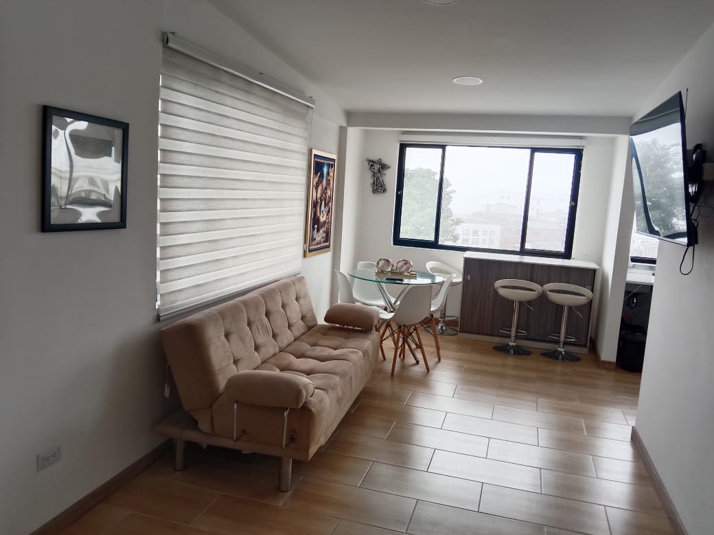 A modern living space features a beige futon and a glass dining table with white chairs. Large windows provide ample natural light, and artwork adorns the walls, contributing to a bright and airy atmosphere.