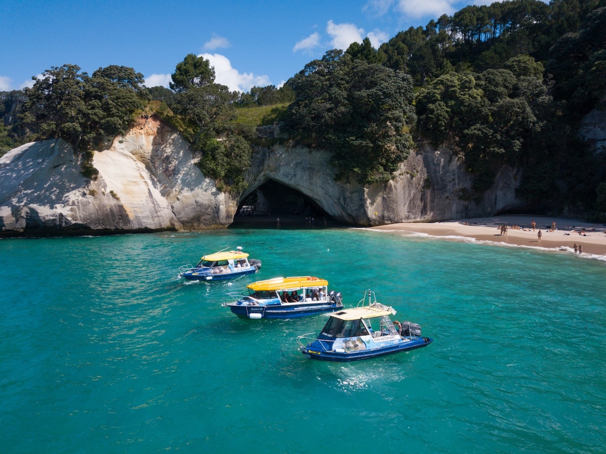 Three boats are anchored in clear turquoise waters near a sandy beach, framed by lush green hills and rocky cliffs. A coastal cave is partially visible, and people can be seen enjoying the beach in the distance under a bright blue sky.