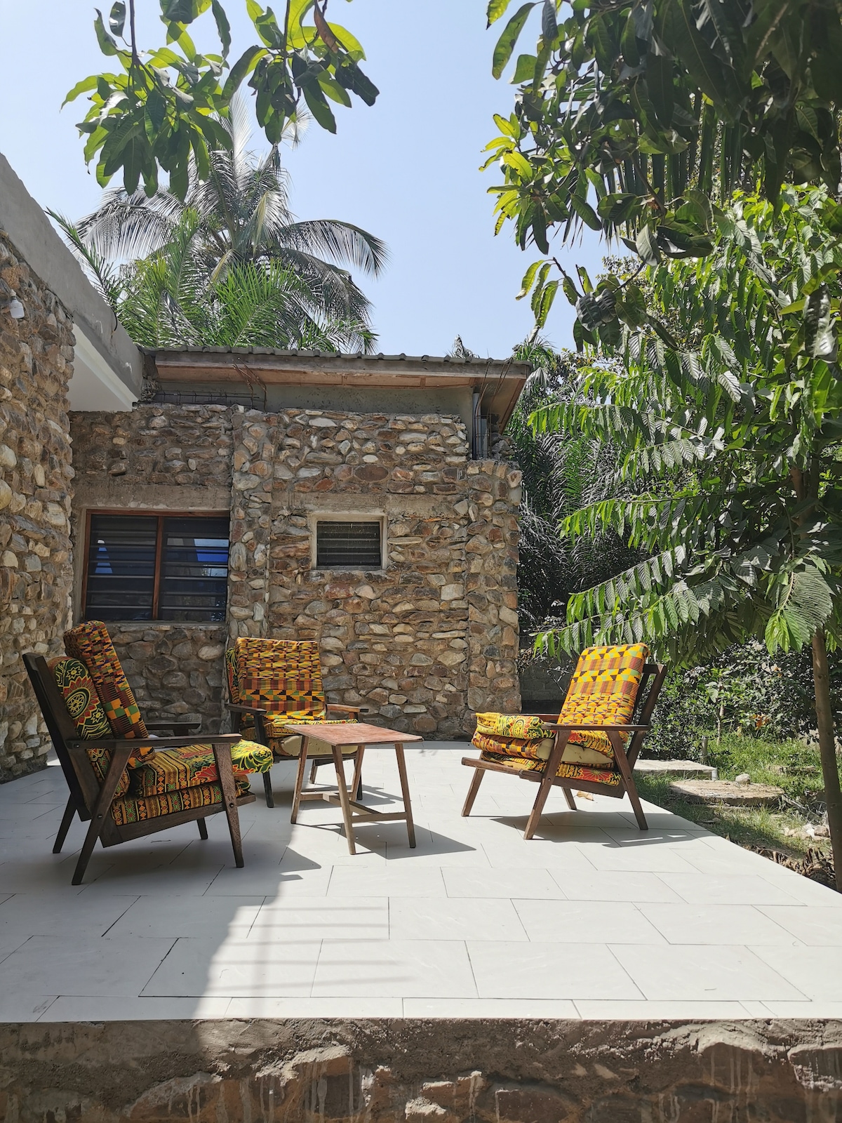An outdoor seating area is featured with four patterned chairs arranged around a small wooden table. The stone wall of the villa is visible in the background, with greenery surrounding the patio. Sunlight is present, creating a bright and open atmosphere.