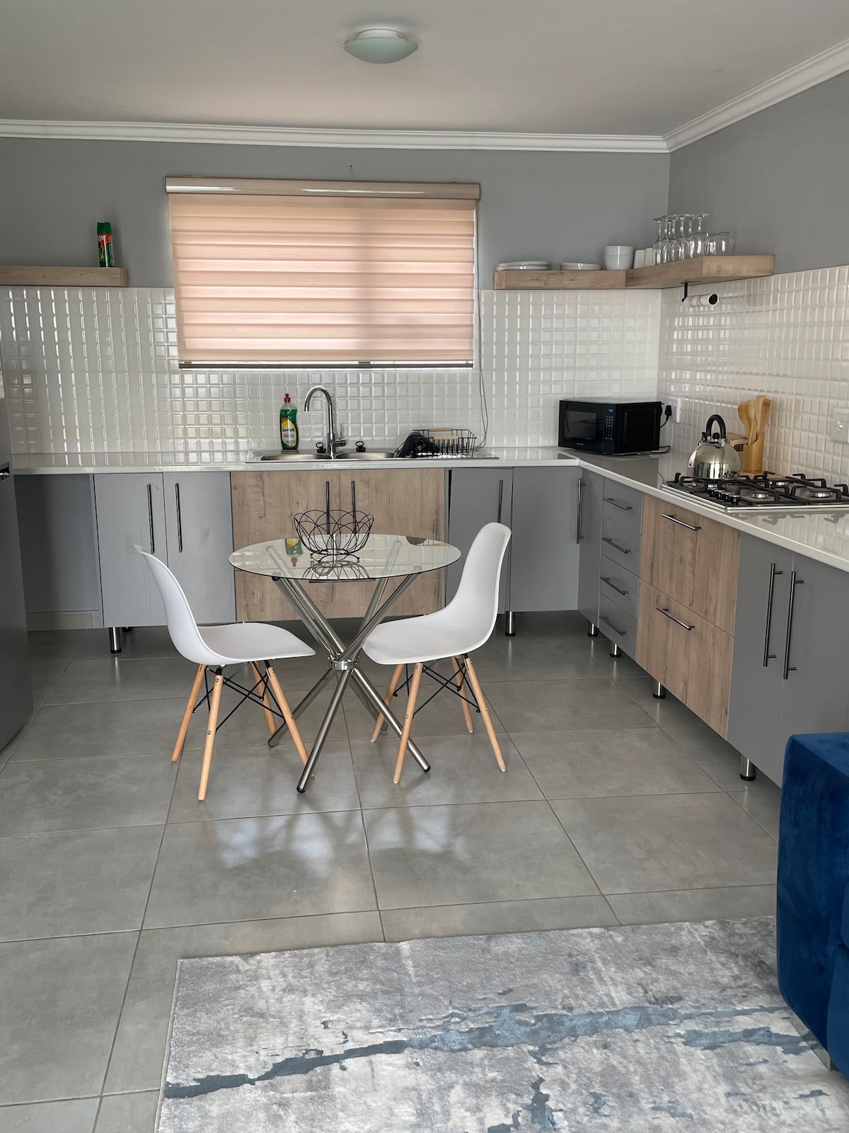 A modern kitchen space features light-colored cabinetry and a neutral tile backsplash. A round glass dining table is paired with two white chairs, creating a minimalist dining area. Natural light filters through a window with a soft blind, illuminating the well-equipped kitchen appliances.