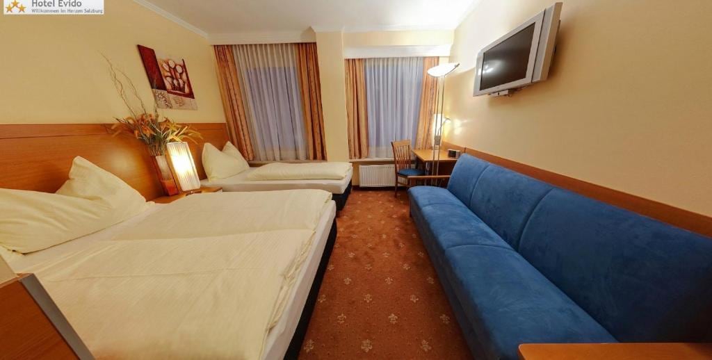 The room features two beds with neutral bedding and a cozy blue sofa positioned against the wall. Natural light fills the space through large windows, partially draped with sheer curtains. A simple desk and chair are also included, along with a wall-mounted television.