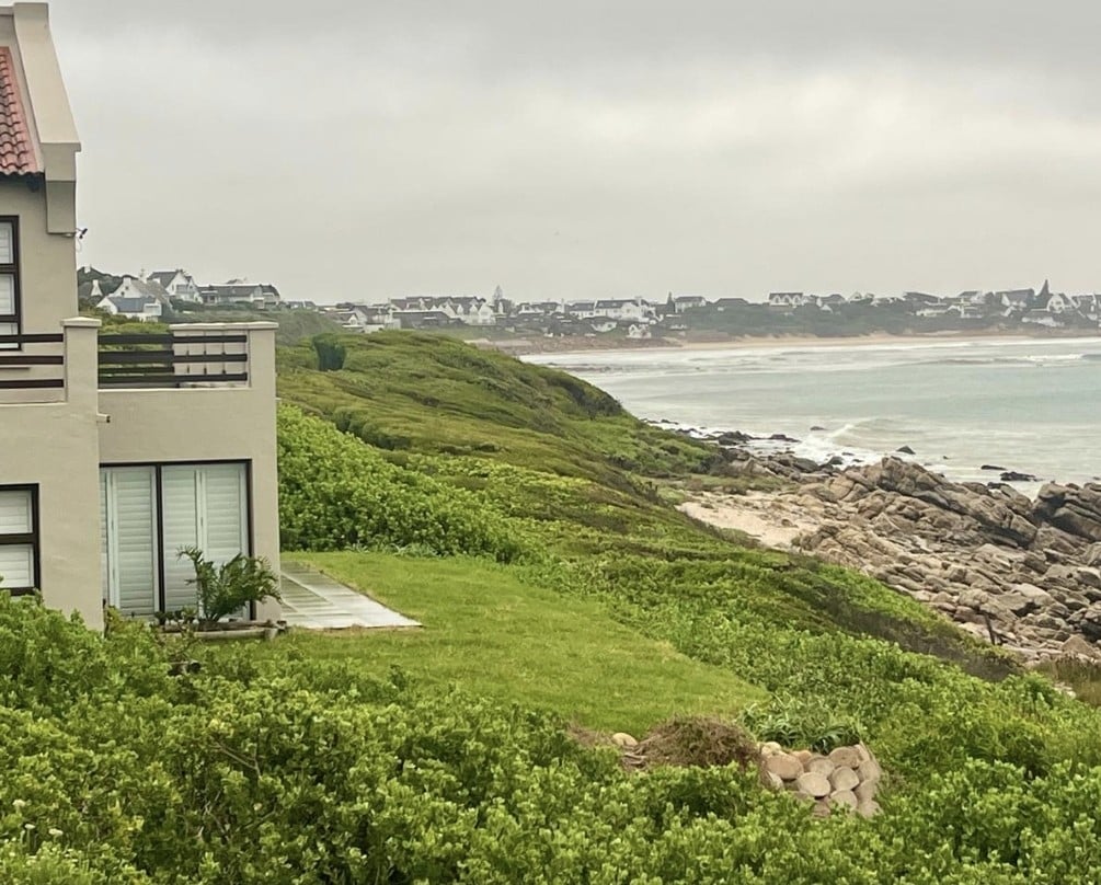 The image captures a coastal view showcasing lush green vegetation adjacent to a well-maintained lawn. A portion of the home is visible, featuring large windows that reflect the landscape. In the distance, the ocean meets the shoreline, framed by gentle waves and rocky outcrops.