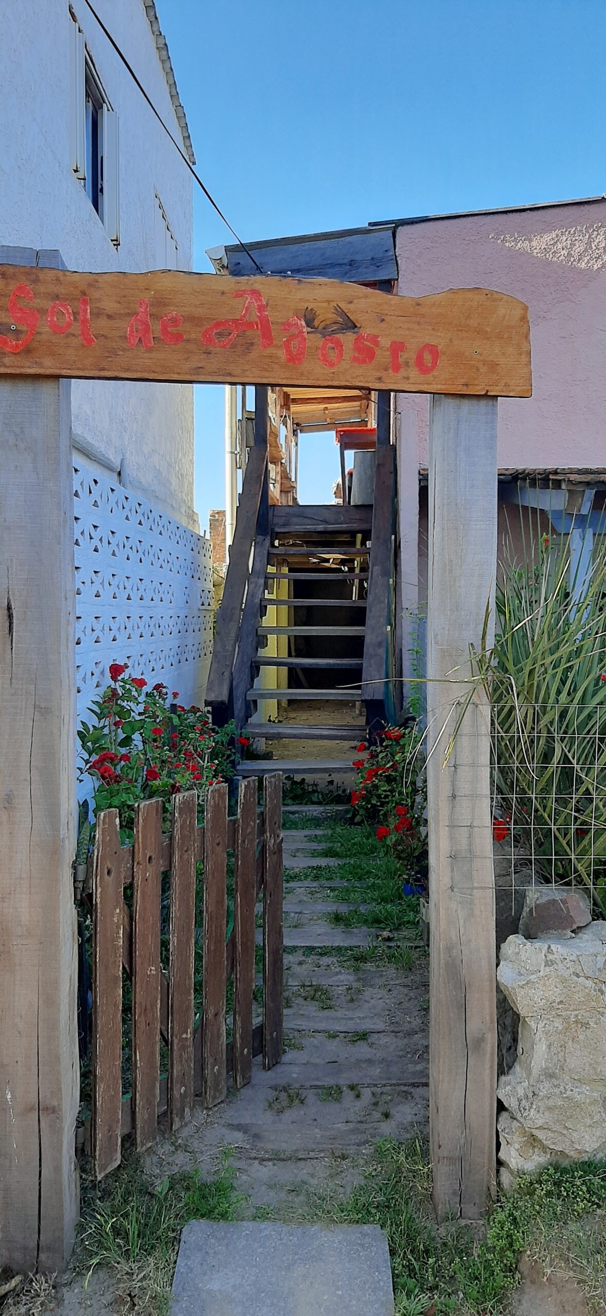 An entrance pathway is framed by a wooden fence and vibrant red flowers. A rustic sign labeled 'Sol de Adosro' is displayed above the steps leading to an upper level. The pathway is lined with greenery, guiding visitors toward the stairs.