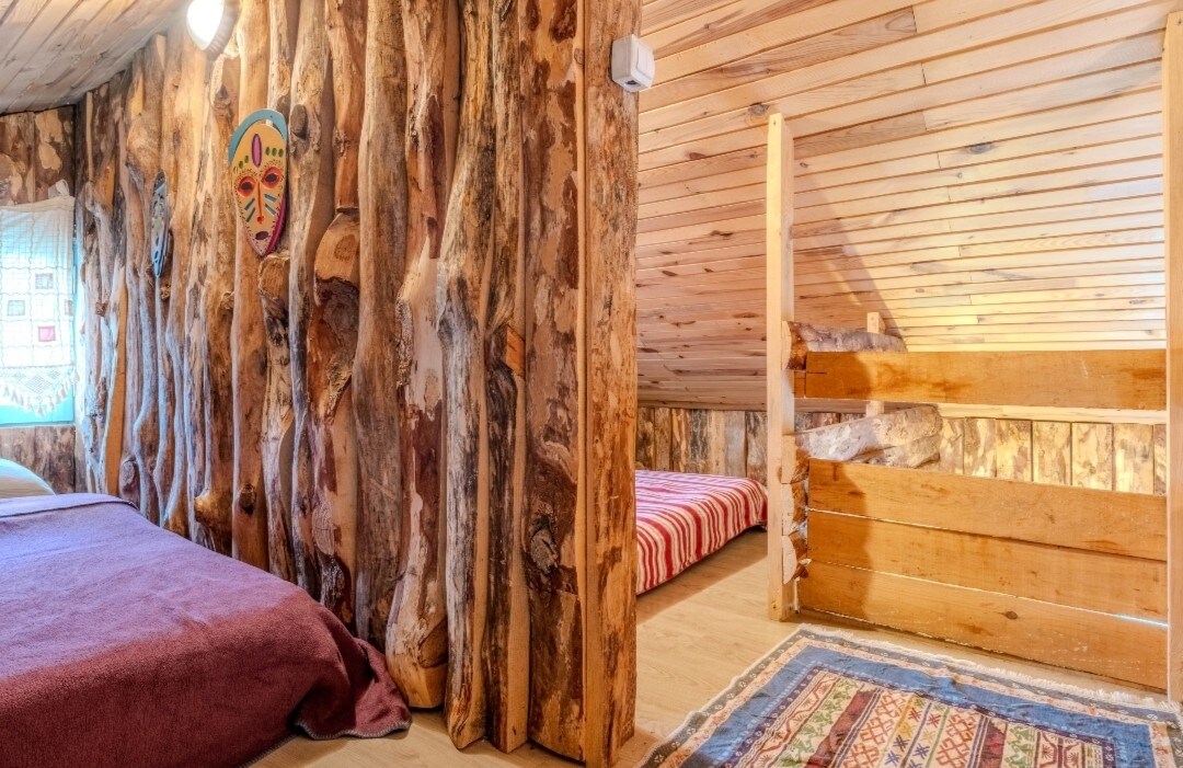 A cozy loft area features wooden walls with natural tree bark details. Two sleeping areas are present, one with a simple patterned blanket and the other with a striped cover. A decorative rug adds warmth to the wood-floored space, illuminated by soft overhead lighting.