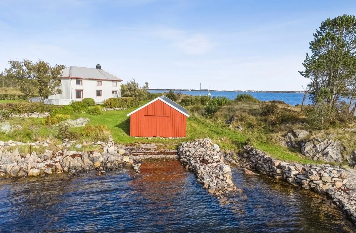 A coastal property is nestled among lush greenery with a red boathouse adjacent to calm waters. The main building, featuring a gray roof, overlooks the sea, creating an inviting outdoor space. Rocky outcrops and plant life punctuate the landscape, enhancing the serene atmosphere.