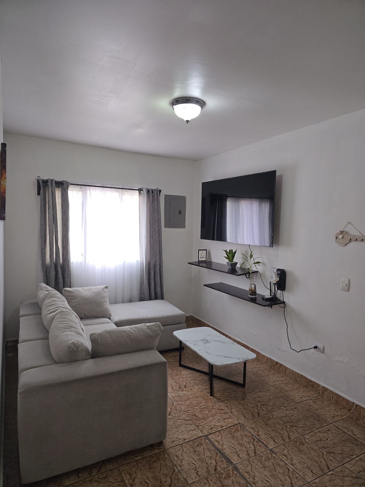 A light and airy living space is filled with natural light from a window draped with sheer curtains. A comfortable sectional sofa is positioned opposite a wall-mounted television. A small coffee table with a marble top is placed in front, complemented by decorative elements on the shelf.