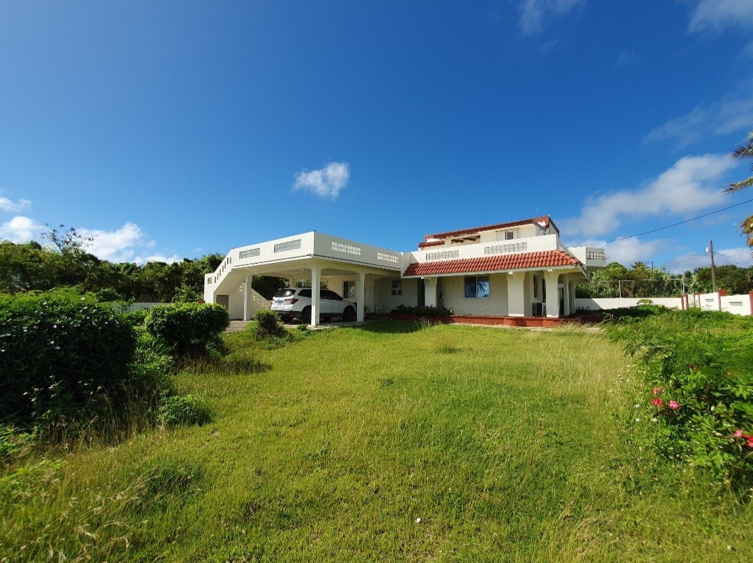 A two-story oceanview house is set within a spacious yard featuring green grass and tropical foliage. The exterior presents a welcoming facade with a red-tiled roof and a covered carport. Bright blue skies enhance the serene atmosphere of this inviting property.