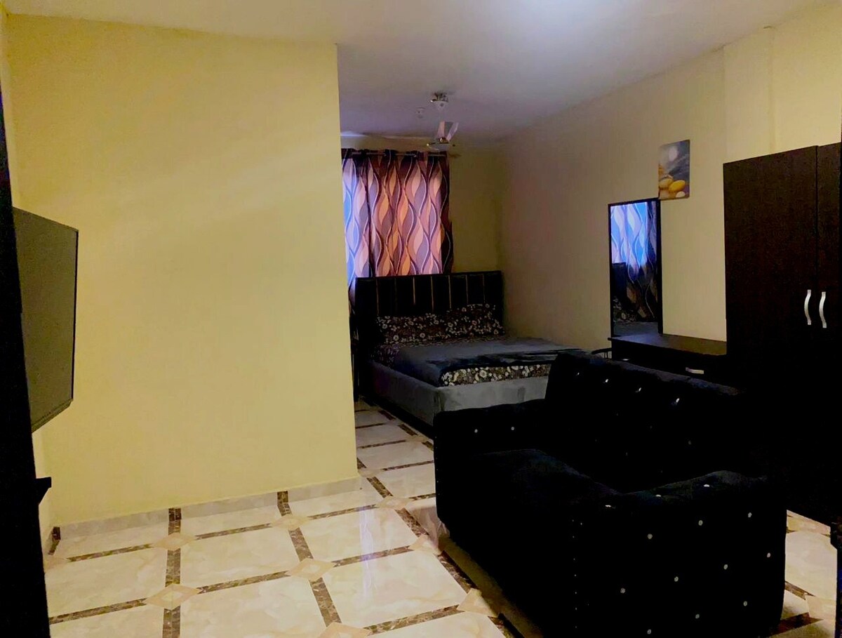 A spacious room features a large bed with a patterned cover placed against one wall. A dark sofa is positioned opposite a simple entertainment unit holding a television. Natural light filters through a window covered by colorful curtains, illuminating the tiled floor.