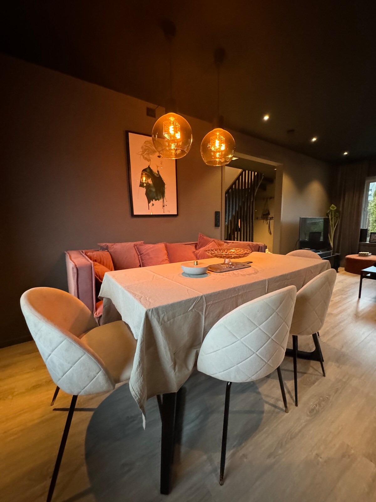 A dining area is presented with a rectangular table set for six, surrounded by plush, light-colored chairs. Two pendant lights provide a warm glow, highlighting the modern decor. A colorful piece of wall art adds interest above the seating space, complementing the contemporary style.