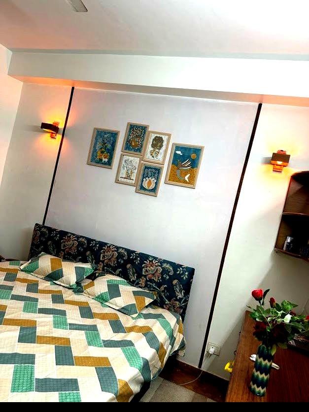 A cozy bedroom is showcased, featuring a spacious bed adorned with a multicolored quilt. The wall above the bed is decorated with framed artworks, while strategically placed wall sconces provide soft lighting. A bedside table and shelf add practicality to the space.