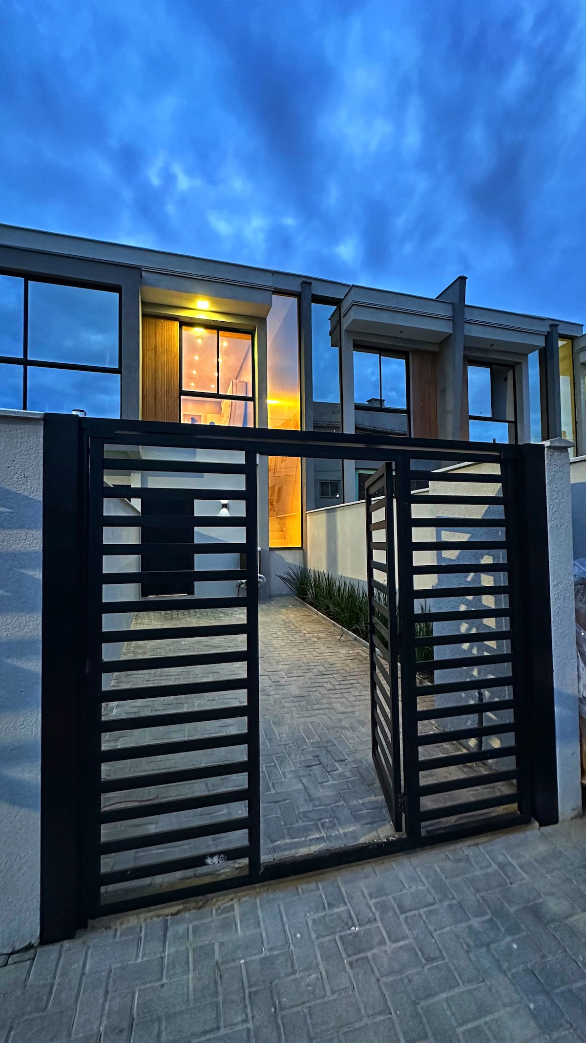 A modern entrance gate with sleek horizontal bars leads to a pathway illuminated by warm lighting. Flanking the walkway are lush green plants, and large windows hint at the inviting interior of the house, framed by dark architectural elements.