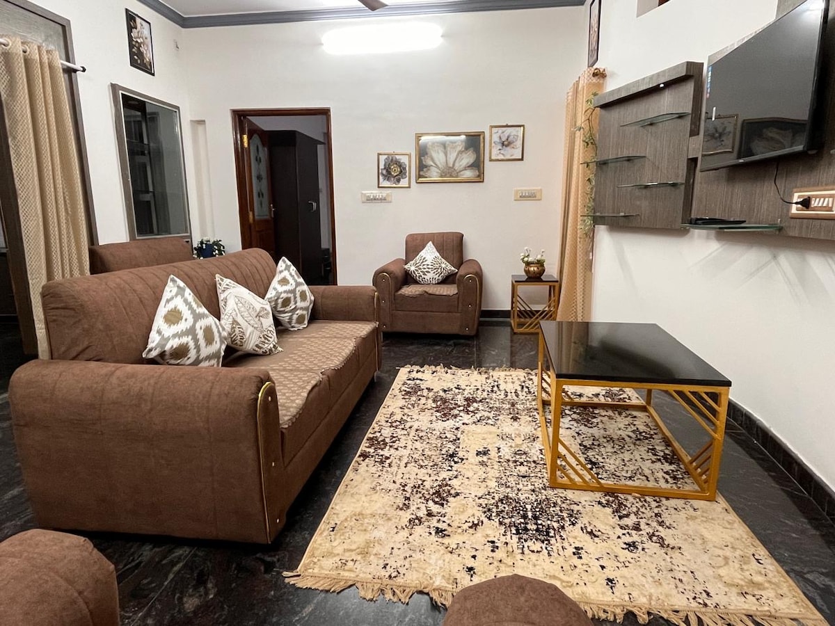 A comfortable living area features a spacious brown sofa with decorative cushions and a matching armchair. A glass-topped coffee table is positioned on a textured area rug, while wall-mounted shelves display decorative items. Soft lighting enhances the room's inviting feel.