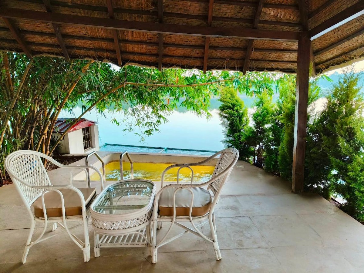 A shaded porch area features a wicker seating arrangement, consisting of two armchairs and a glass-topped table. Lush greenery surrounds the space, and the view of the water can be seen beyond a gentle slope, providing a serene setting for relaxation.