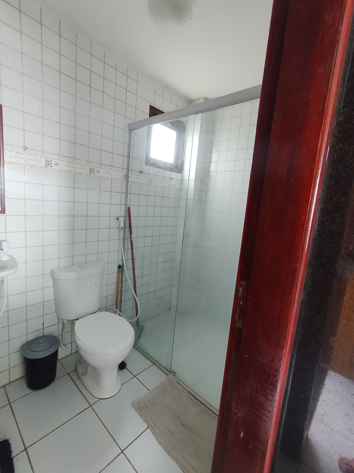 The bathroom features white tiled walls and a glass shower enclosure, allowing for natural light to enter. A toilet and a small waste bin are positioned nearby, with a bath mat lying on the tiled floor. Basic amenities may be present for guest convenience.