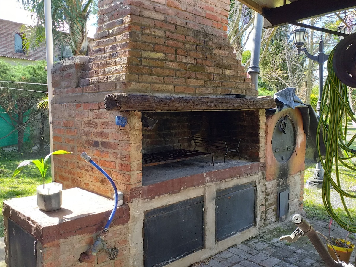 A traditional outdoor grill area is depicted, featuring a stone brick construction with a spacious grilling space above and a side area for preparation. Nearby, a small potted plant is visible, along with garden hoses and tools, indicating a functional outdoor cooking area.