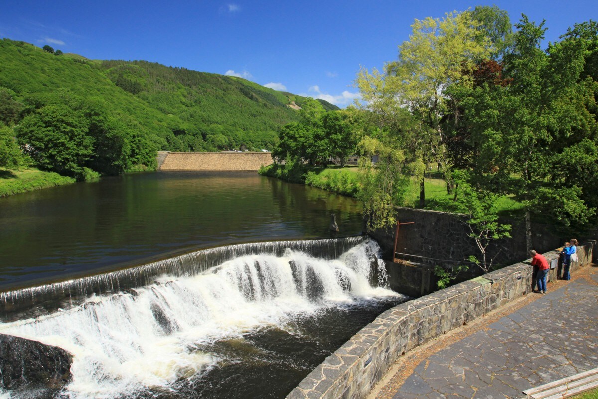A serene river scene captures a waterfall cascading over a stone wall into a calm body of water. Lush green hills line the background, framed by a clear blue sky. A few individuals can be seen near the water's edge, enjoying the natural landscape.