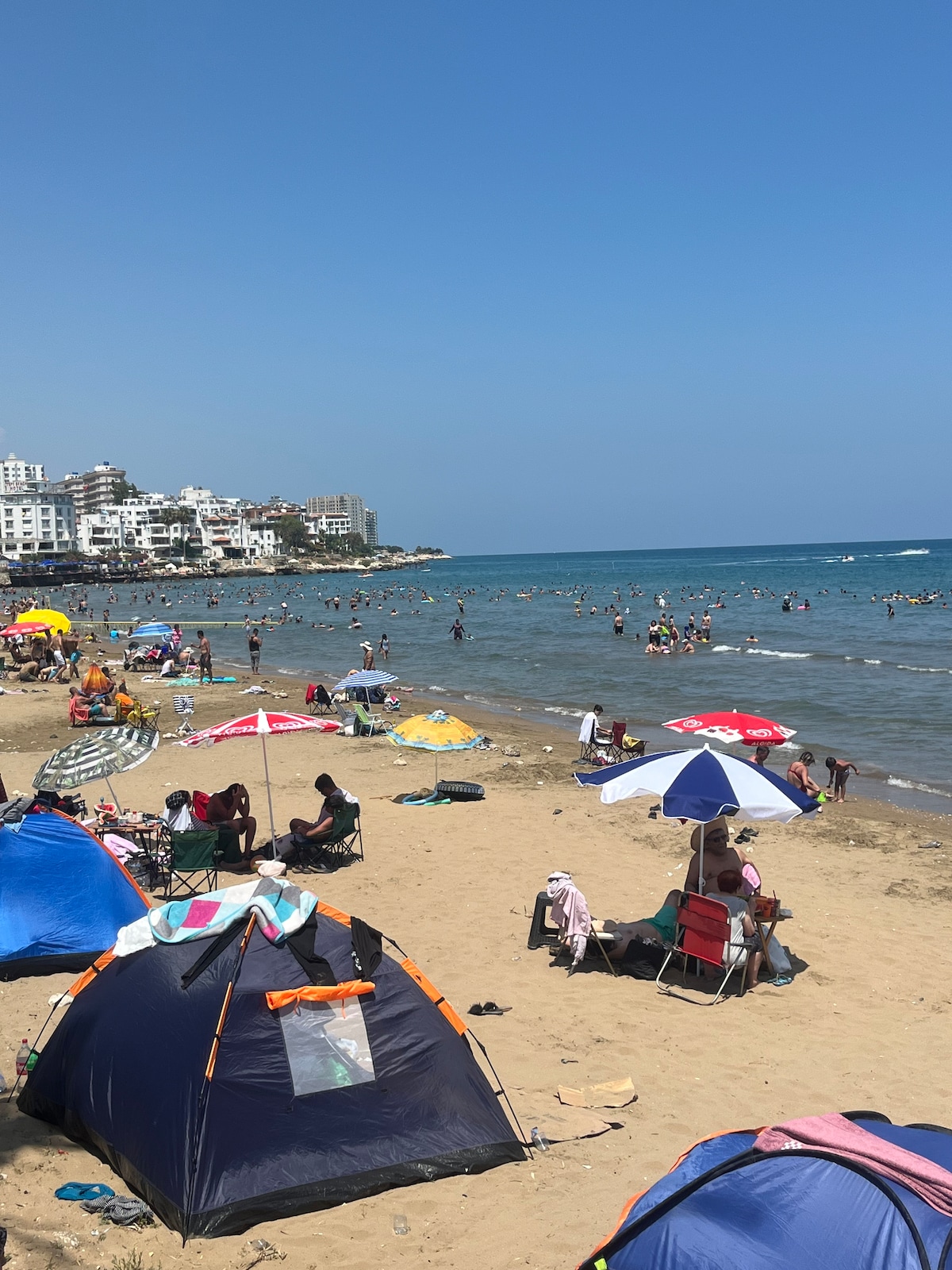 The beach scene depicts many colorful umbrellas and tents scattered across the sandy shore. People are seen enjoying the water and sunbathing, while a collection of buildings borders the beach in the background, contributing to the vibrant atmosphere.