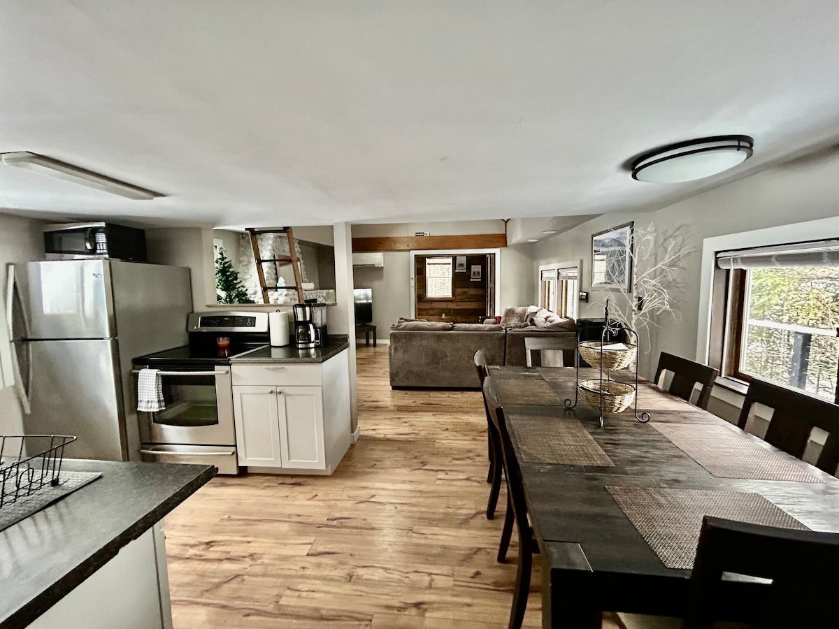 An open concept living space features a dining table with ample seating, a kitchen equipped with modern appliances, and a comfortable living area with a sofa. Large windows allow natural light to fill the room, enhancing the inviting atmosphere.